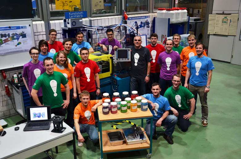Das Bild zeigt eine Gruppe von Studierenden und möglicherweise Dozenten in einer technischen Werkstatt oder einem Labor. Die Gruppe trägt bunte T-Shirts in verschiedenen Farben, auf denen ein Symbol abgebildet ist, das wie eine Glühbirne aussieht, was auf kreative Ideen oder Innovation hinweisen könnte. Im Hintergrund ist eine „Kunststoff-Lernfabrik“ zu sehen, und eine Maschine, vermutlich eine Spritzgussmaschine, steht im Zentrum der Szene. Auf einem Tisch im Vordergrund sind mehrere farbige Behälter mit Kunststoffgranulat zu sehen, was darauf hindeutet, dass die Gruppe möglicherweise an einem Projekt im Bereich Kunststoffverarbeitung arbeitet. Die Atmosphäre ist freundlich und kooperativ, und das Bild vermittelt den Eindruck einer praxisorientierten Lernumgebung, in der die Gruppe an einem gemeinsamen Projekt im Bereich Maschinenbau oder Kunststofftechnik arbeitet.
