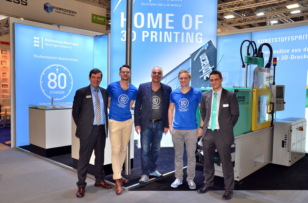 Das Bild zeigt eine Gruppe von fünf Personen, die vor einem Ausstellungsstand auf einer Messe stehen, die sich um 3D-Druck-Technologie dreht. Der Stand trägt den Schriftzug „Home of 3D Printing“ und zeigt den Bezug zur Hochschule Reutlingen, speziell dem Studienbereich Maschinenbau. Zwei der Männer tragen blaue T-Shirts mit dem Aufdruck „80 Stunden Challenge“, was auf ein Projekt oder eine besondere Veranstaltung hinweisen könnte. Die anderen drei Männer tragen Anzüge, was auf eine Mischung aus Studierenden und Fachkräften hinweist. Im Hintergrund ist eine Maschine zu sehen, die wahrscheinlich Teil einer Demonstration der 3D-Druck- oder Kunststoffspritzguss-Technologie ist. Die Atmosphäre ist professionell und auf Technologie und Innovation fokussiert, was darauf hinweist, dass das Team seine Entwicklungen oder Forschungsprojekte im Bereich des 3D-Drucks präsentiert.