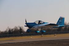 Das Bild zeigt ein kleines, blau-weißes Propellerflugzeug, das gerade vom Boden abhebt oder landet. Das Flugzeug befindet sich in der Nähe einer Start- oder Landebahn, und im Hintergrund ist eine Landschaft mit Bäumen zu sehen. Die Aufnahme wurde wahrscheinlich auf einem Flugplatz gemacht. Das Flugzeug trägt Markierungen auf dem Heck und wirkt wie ein leichtes Sport- oder Trainingsflugzeug. Der Propeller ist in Bewegung, was auf den aktiven Flugzustand hindeutet. Die Szene vermittelt Dynamik und Flugbegeisterung, möglicherweise im Rahmen eines Projekts oder eines Trainingsfluges.